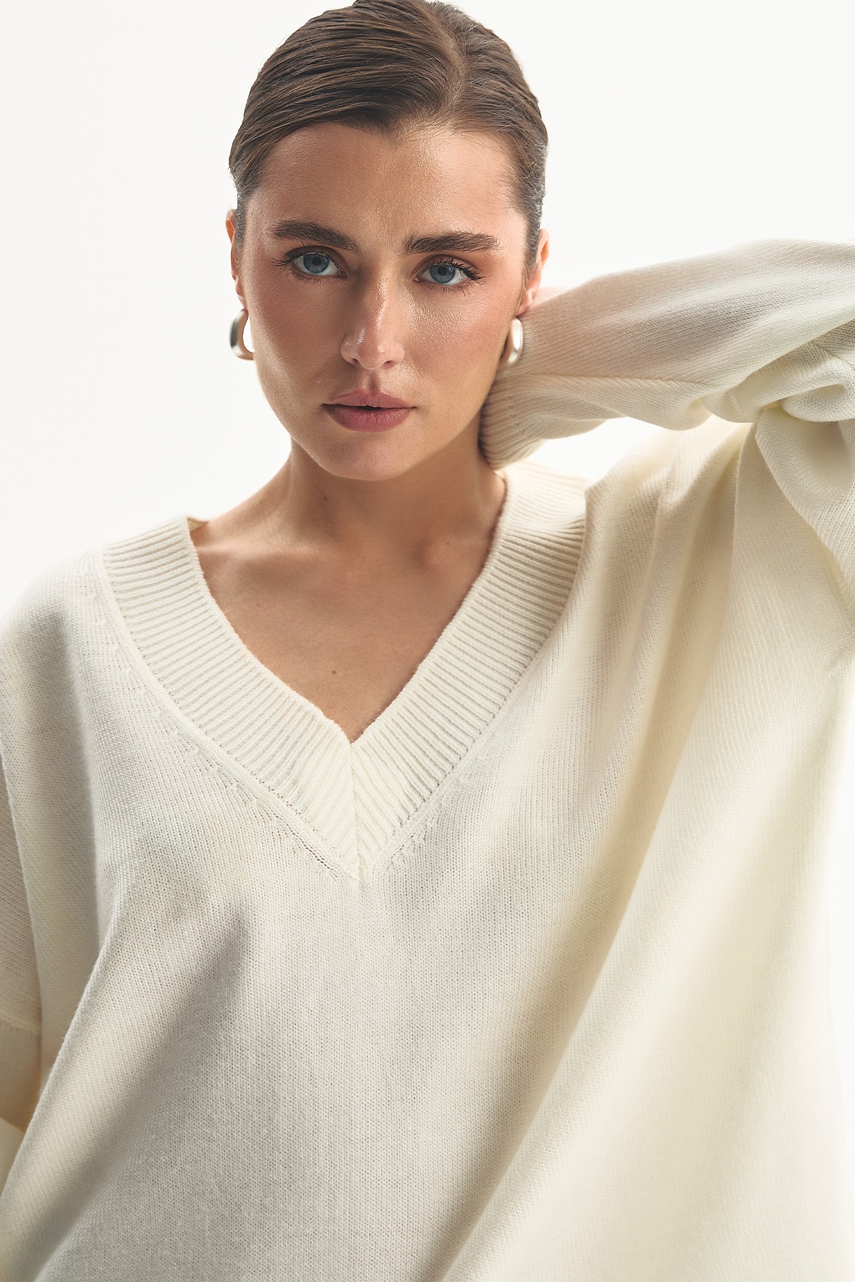 Молочний светр oversize з глибоким V-подібним вирізом і резинкою по краю, рука закинута за шию
